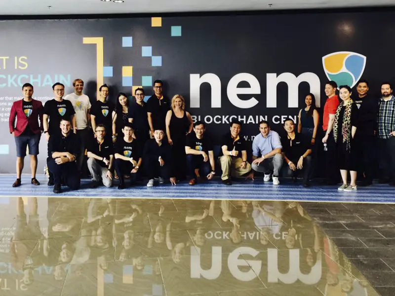 A group photo of people in front of a wall with the NEM Blockchain Centre logo, showcasing team members and attendees.