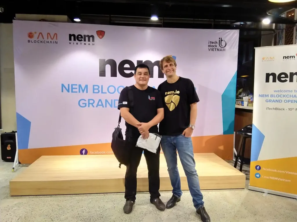 Two individuals stand in front of a backdrop that reads "NEM Blockchain Grand Opening" with logos for NEM Vietnam, AM Blockchain, and iTech Black Vietnam. The backdrop and banners prominently display the NEM logo, illustrating the grand opening event.