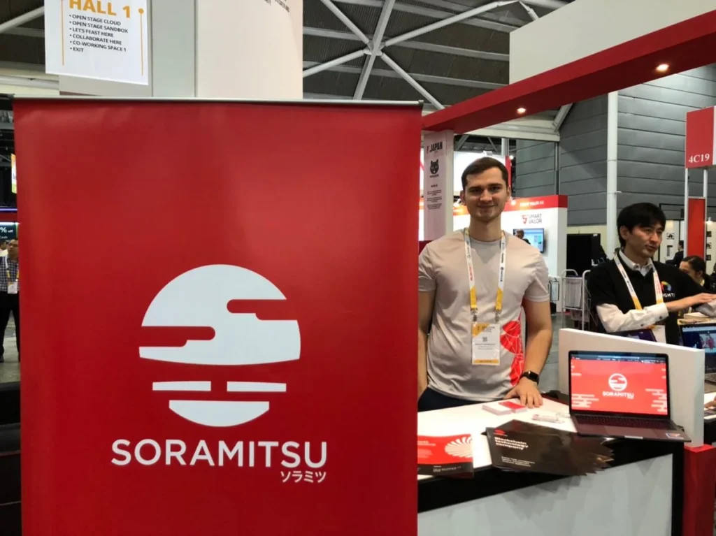 An exhibitor at a trade show booth featuring the Soramitsu logo and branding materials.