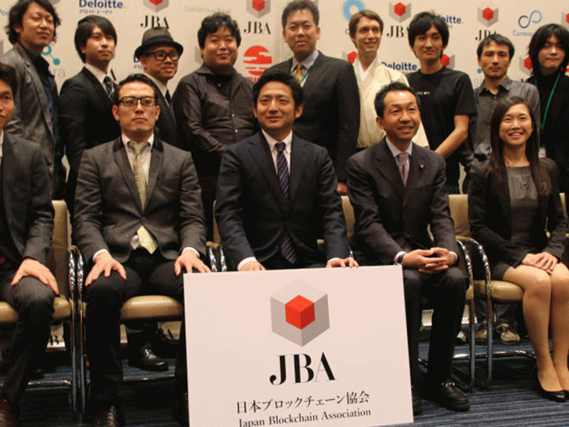 Members of the Japan Blockchain Association (JBA) posing for a group photo in front of a backdrop featuring sponsor logos, with a prominent JBA sign in the foreground.